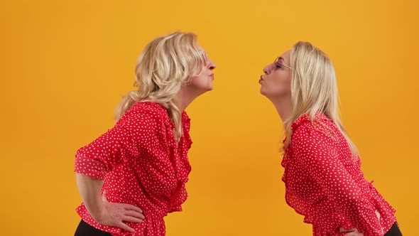 Two Blonde European Women Leaning Forward to Kiss Medium Shot Orange Background Studio Shot
