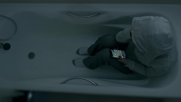 A Young Woman in a Hood Sits in the Bathroom and Looks at the Screen of Her Phone