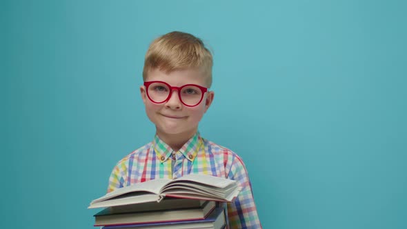 Pretty Schoolboy in Eyeglasses Reading Book and Smiling Looking at Camera Standing on Blue