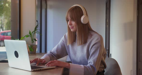 Cheerful Young Woman in Headphones Listening to Music Using Laptop in Cafe