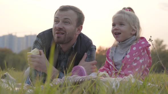 Portrait of Caucasian Father and His Little Blonde Daughter Sitting on the Meadow and Eating Apples