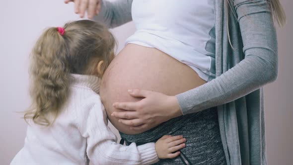 Little Kid Hugs Pregnant Mommy Showing Tummy at White Wall