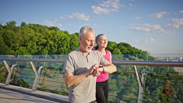 Active Elderly Man and Woman Jog on Bridge Against River