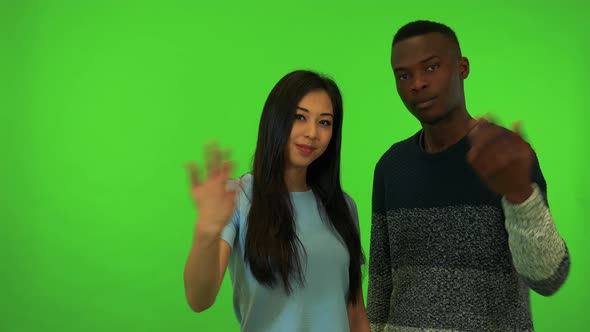 A Young Asian Woman and a Black Man Wave at the Camera in a Gesture of Invitation - Green Screen