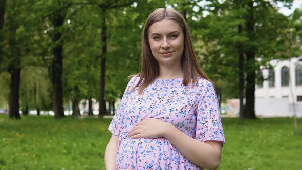 A Pregnant Woman in a Summer Dress with a Floral Print Walks in the Park and Breathes Fresh Air