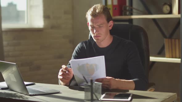 Young Man of Business Sitting in the Office at the Table Near Notebook and Working at Documents