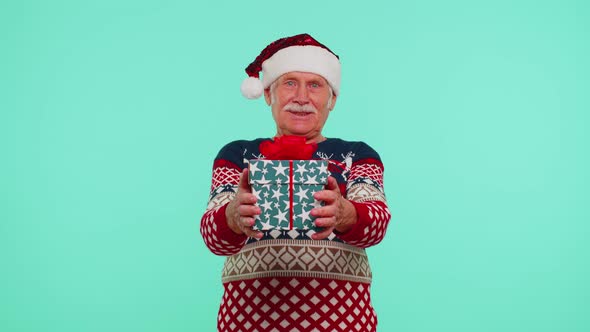 Grandfather in Santa Sweater Presenting One Christmas Gift Box Stretches Out His Hands to Camera