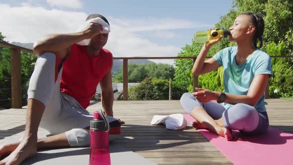 Tired biracial couple on terrace drinking water, resting after exercises and talking