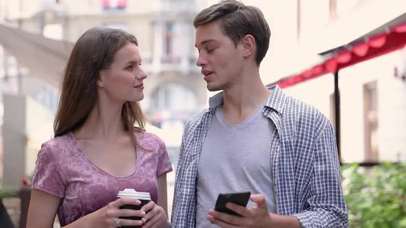 Happy Couple Using Mobile Phone And Drinking Coffee Outdoors