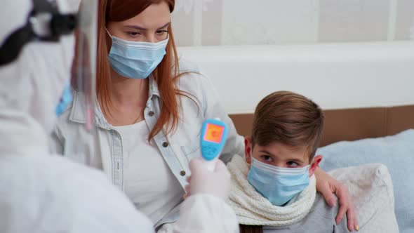 Nurse Health Worker in Protective Suit Treats Small Male Patient and Measures High Body Temperature