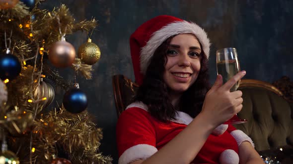 Young Woman in Santa Claus Costume with Glass of Champagne Sitting on Chair Near Christmas Tree