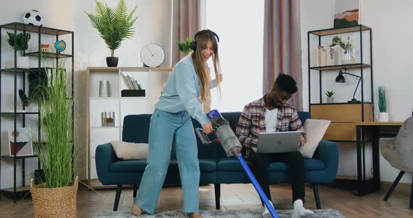 Girl in Headphones Cleaning Carpet with Vacuum Cleaner while Her Black-Skinned boyfriend