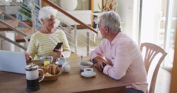 Senior caucasian couple talking to each other using smartphone and laptop having breakfast together