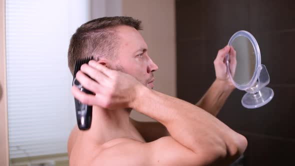 A young man cuts himself, using reflection mirrors and a hair clipper