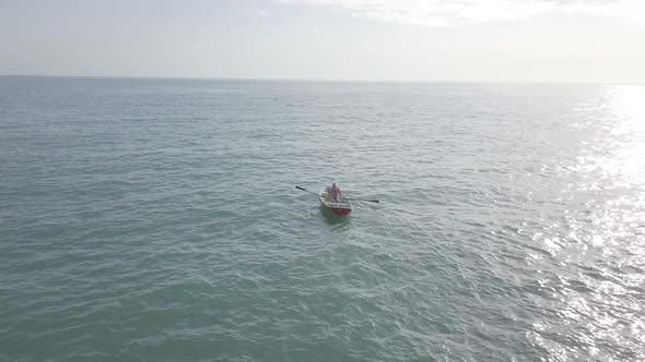 Aerial view of fisherman rowing oars in small fishing boat floating in open sea. Sunlight on water