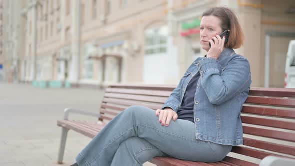 Angry Old Woman Talking on Phone While Sitting Outdoor on Bench