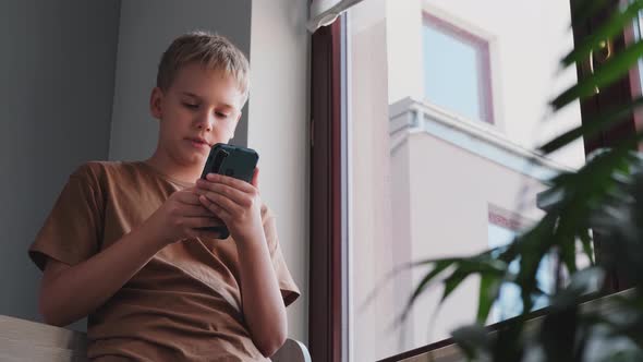 A Teenager Uses a Smartphone Phone While Standing Near the Window in His Room