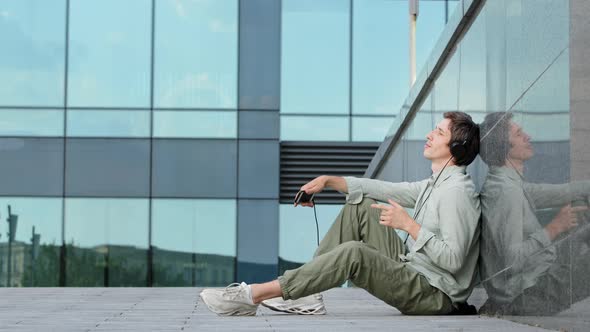 Happy Millennial Guy in Earphones Sitting on Ground in Front of Modern Glass Building Enjoy Rhythmic