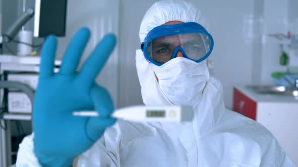 Doctor in a Hazmat Suit Is Holding a Thermometer Showing Fever