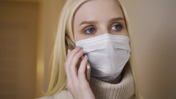 Young Girl in Medical Protective Mask Sits at the Table in Cafe and Talks on Smartphone