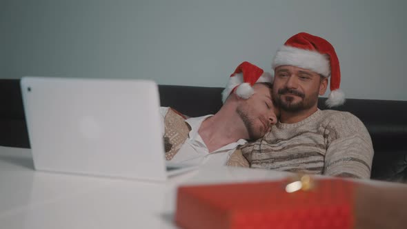 Happy Gay Male Couple with Santa Claus Hats Cuddling and Watching Movie on the Christmas Eve