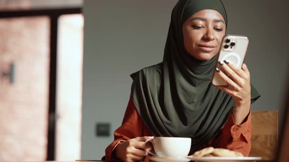 Smiling Muslim woman texting by phone with cup of tea