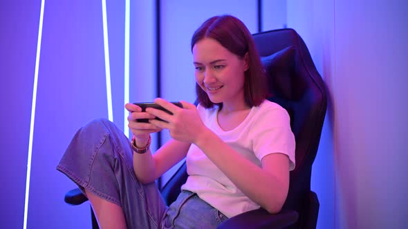 Young Girl Playing Mobile Online Game on a Smart Phone While Sitting on Gaming Chair in Colorful