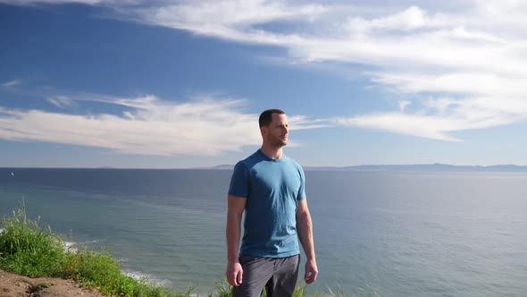 A fit strong man standing proud on the edge of an ocean cliff nature hiking trail with an epic beach