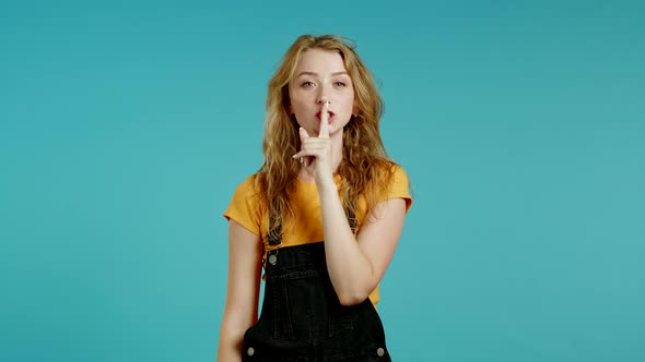 Smiling Woman with Blonde Hair Holding Finger on Her Lips Over Blue Background. Gesture of Shhh