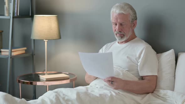 Old Man Celebrating Success on Documents in Bed