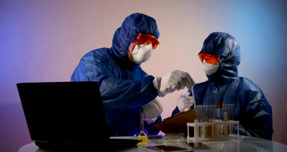 People in Protective Clothing with Hoods, Respirators, Glasses and Gloves Sit at a Table with a