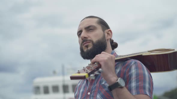 Close-up of Bearded Handsome Man with Guitar Looking Away and Thinking. Young Caucasian Musician