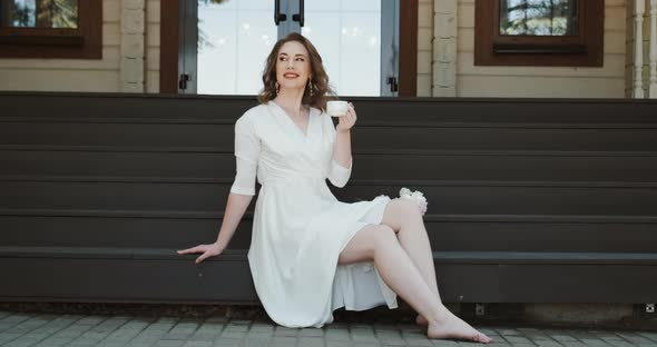 Attractive Woman in White Dress Drinking Tea or Coffee on Terrace of Her House