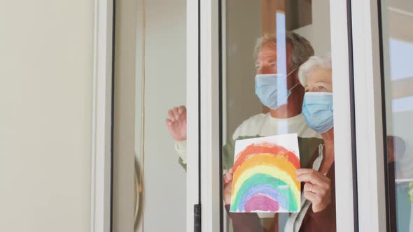 Senior caucasian couple wearing face masks holding rainbow painting against the window at home