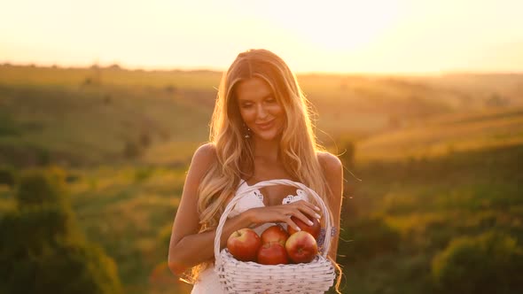 Beautiful sexy blonde girl in white dress posing in a field at sunset with a basket of fruit	
