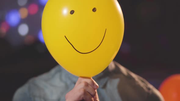 Portrait of a Confident Young Man in Birthday Hat Removing a Balloon Painted with Funny