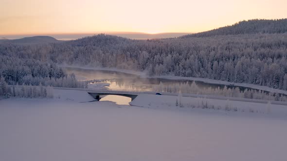 Aerial view of fast car car driving over ice lake bridge under glowing sunrise snowy woodland mounta