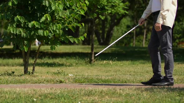 Man With Visual Impairment Walking Along Path in Park Groping His Way With Cane