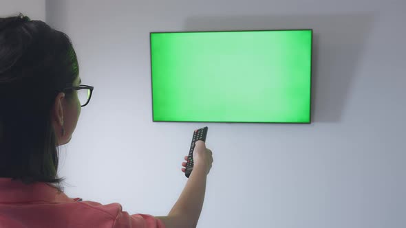 Girl with Glasses Sits on a Sofa in Front of the TV and Switches Channels with a Remote Control