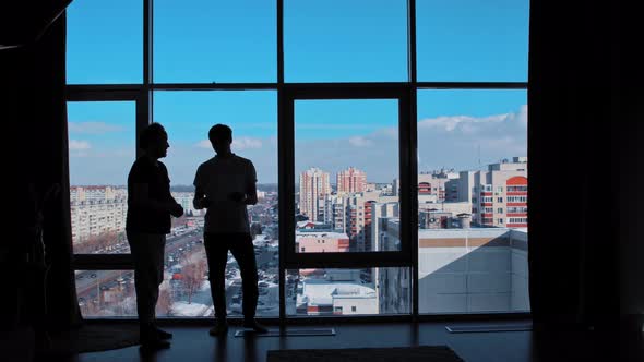 Two Businessmen Stand in an Office with Paronamy Windows and Discuss Papers