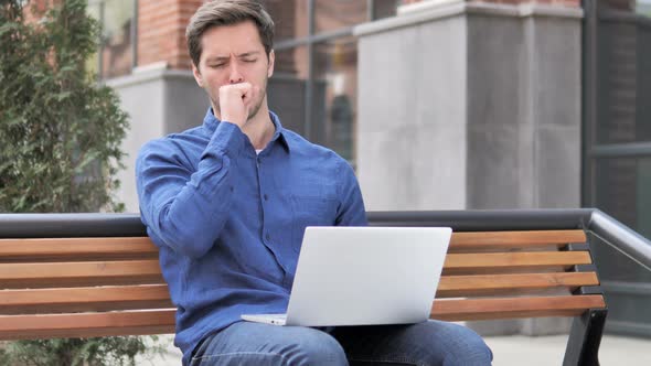 Sitting Outdoor Young Man Coughing while Working on Laptop