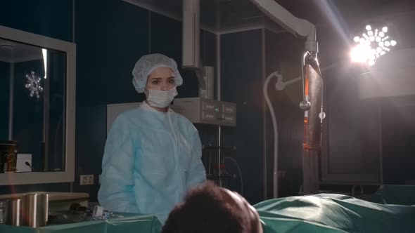 Female Surgeon in Sterile Gloves and a Robe Approaches the Patient on the Operating Table