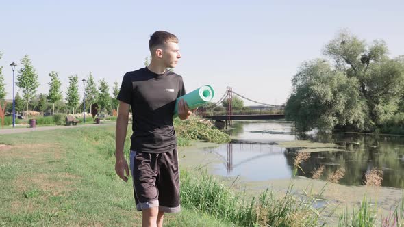 Young man walking along river in park with yoga mat in hands