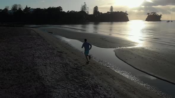 Aerial follow shot of silhouetted young man running on a beach in Auckland, New Zealand