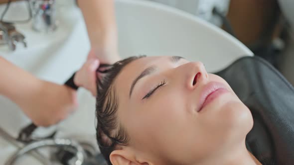 Caucasian young woman lying down and close eye on salon washing bed getting hair washed by stylist.