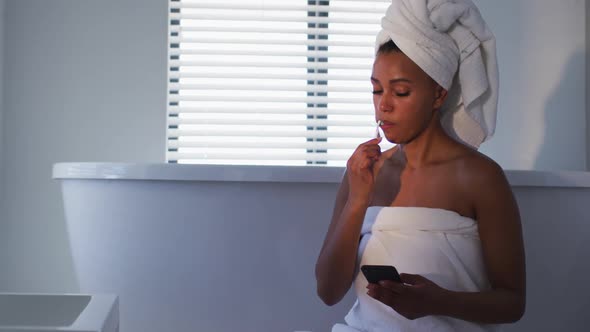 African american woman using smartphone while brushing her teeth in the bathroom