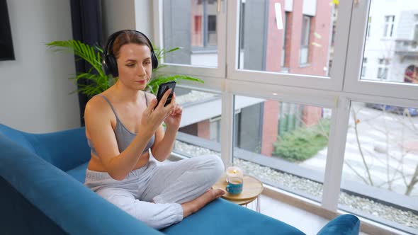 Woman in Wireless Headphones Listening to Music and Using Mobile Apps on Smartphone Sitting on Sofa