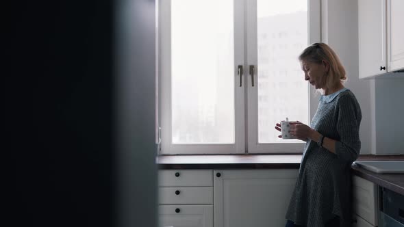 Lonely Elderly Woman Drinking Coffee Standing at Her Kitchen