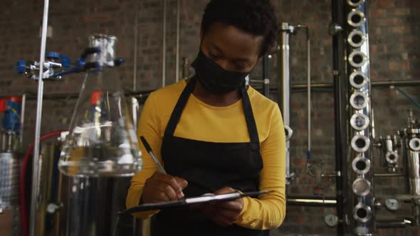 African american woman in face mask working at distillery checking equipment, writing on clipboard