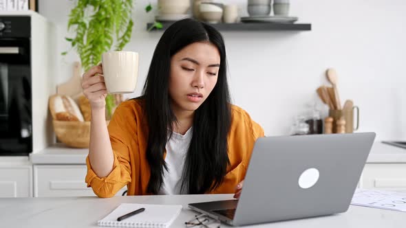 A Sleepy Asian Girl Freelancer or a Student Sits at Home in the Kitchen at a Table with Her Eyes